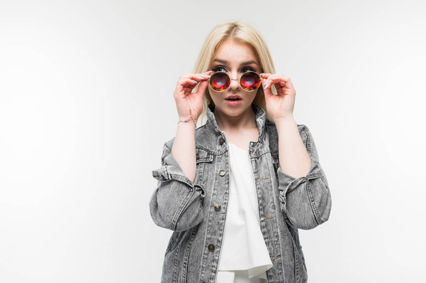 portrait of young charming joyful blonde girl in jeans jacket possing and gesticulating in round sunglasses on background - Photo, Image