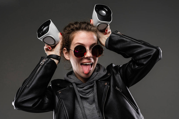 fashionable young woman in leather jacket and sunglasses posing with loudspeakers against dark background - 写真・画像