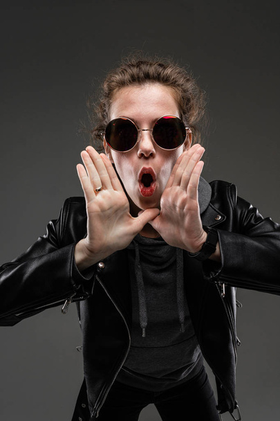 модна молода жінка позує в шкіряній куртці і сонцезахисних окулярах проти сірого
  - Фото, зображення