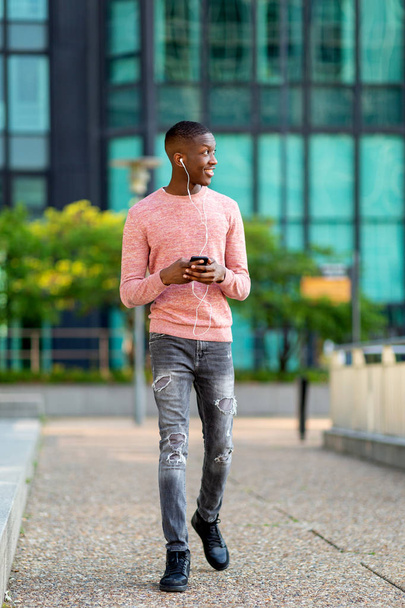 Full length portrait young black man walking with mobile phone and earphones - Foto, imagen