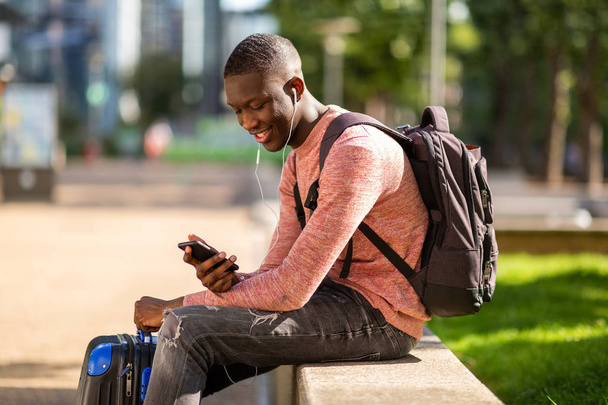 Бічний портрет молодого африканця, що сидить на вулиці з сумками для подорожей та мобільним телефоном
 - Фото, зображення