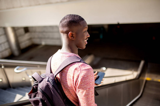 Portrait from behind of young black man going down escalator with mobile phone - Fotó, kép