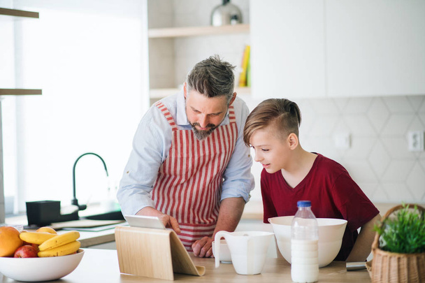Mature father and small son with tablet indoors in kitchen, making pancakes. - Foto, Bild