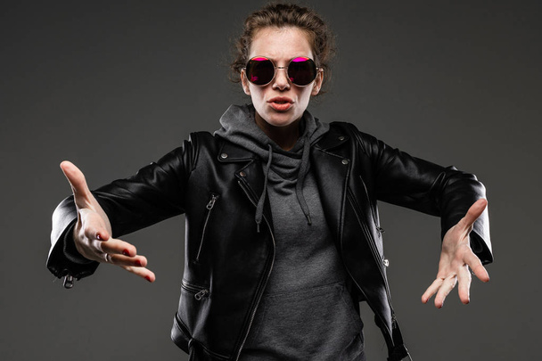 fashionable young woman posing in leather jacket and sunglasses against dark background - Photo, image
