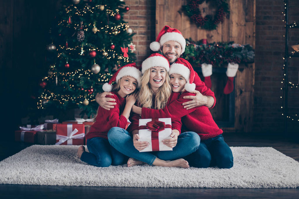 Full four members family dad mom sister brother giving surprise giftbox spending x-mas eve sitting floor near newyear tree indoors wear santa caps and red sweaters - Foto, imagen