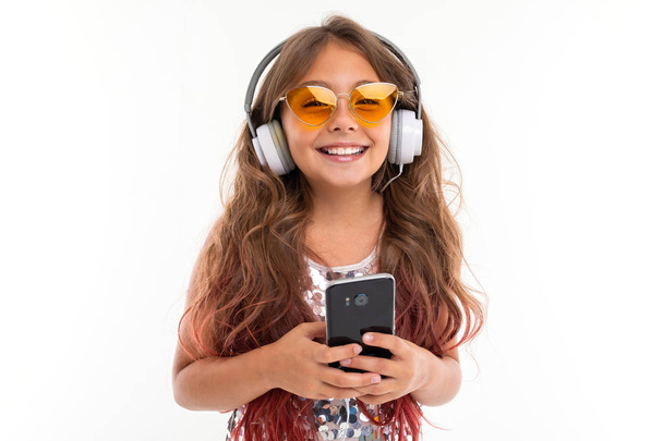 Gelukkig glimlachend meisje in glitterjurk en gele zonnebril, met grote oortelefoons luisteren naar muziek en houden mobiele telefoon geïsoleerd - Foto, afbeelding
