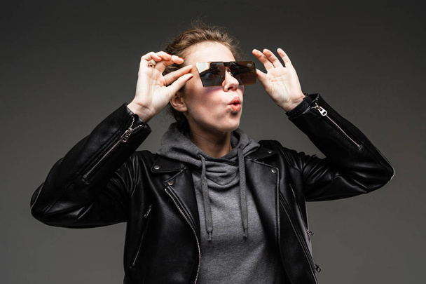 mujer joven de moda posando en chaqueta de cuero y gafas de sol contra gris
  - Foto, Imagen