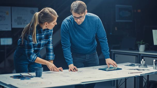 In the Dark Industrial Design Engineering Facility Male and Female Engineers Talk and Work on a Blueprints Using Digital Tablet and Conference Table. On the Desktop Drawings and Engine Components - 写真・画像