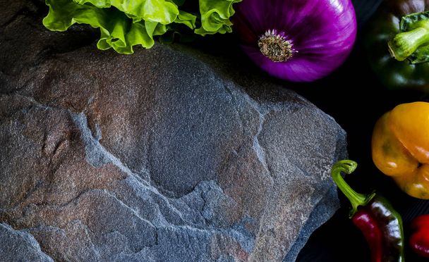 A wild multi-colored stone with a place for text lies on a black table next to fresh blue onions, yellow peppers, chili and lettuce - Foto, imagen