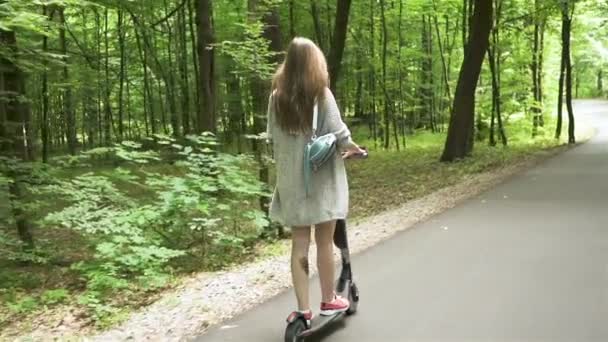 Movimento lento. Rapariga em casaco cardigan montando scooter elétrico na área do parque da cidade. Disparo firme
 - Filmagem, Vídeo