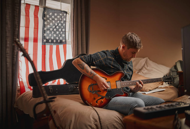 Young guitarist man at home sitting on bed in front of window with usa flag playing guitar - Photo, image
