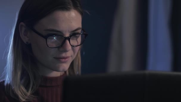 working woman, beautiful female works at a computer in evening, smiles, prints text and looks into monitor with glasses to protect her eyesight - Кадри, відео