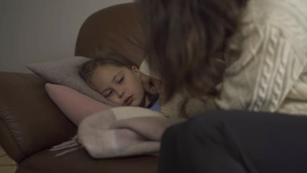 Close-up of sick caucasian girl lying under blanket at home. Sad child has fever. Young mother checking daughters temperature on thermometer. Concept of health, illness, sickness, cold, treatment - Video, Çekim