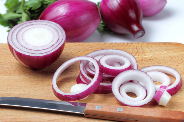 Sliced rings of red onion on a cutting board made of wood, in the background whole heads of onions and fresh herbs, white background. - Foto, afbeelding