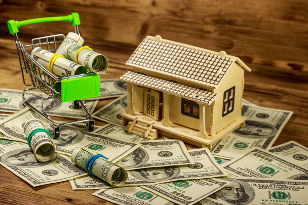 Hausmodell und kleiner Einkaufswagen mit Dollarrollen auf einem Stapel von Hundert-Dollar-Scheinen auf Holzgrund. Immobilieninvestitionen, Eigenheimdarlehen, Hypothekendarlehen, Immobilienkonzept - Foto, Bild