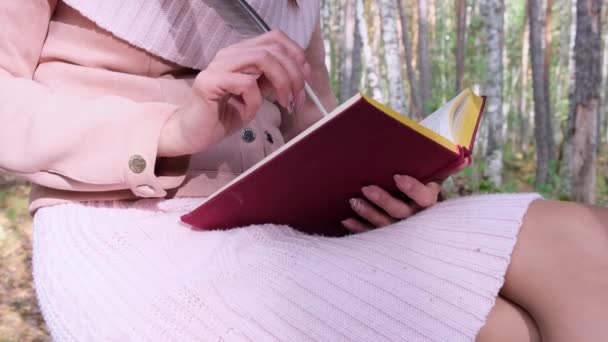 秋の森の女の子のクローズアップショット,女性の手は本にペンで書きます.女性は秋の森の木の近くに座って、彼女の手に本を持っています. - 映像、動画