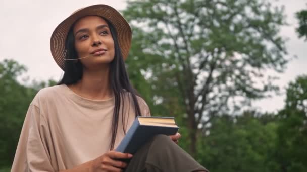 Thoughtful cute young brunette asian woman in hat holding a book and chewing a straw while sitting in the park - Video