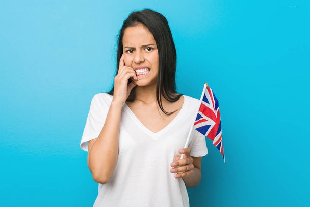 Birleşik Krallık bayrağı tutan genç İspanyol kadın tırnaklarını ısıran, gergin ve çok endişeli. - Fotoğraf, Görsel