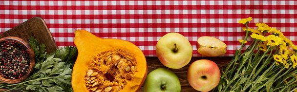 top view of pumpkin, peppercorn, yellow flowers and apples on wooden table with red napkin, panoramic shot - Photo, Image