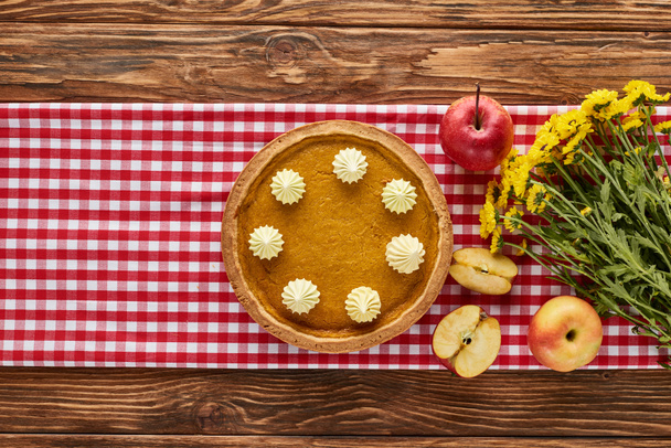 vue du dessus de tarte à la citrouille, pommes et fleurs jaunes servis sur table en bois avec serviette à carreaux rouge
 - Photo, image