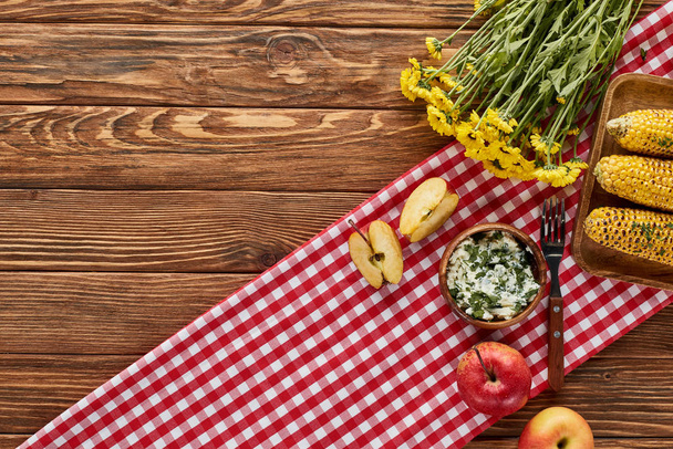 Kırmızı peçetedeki ahşap masada servis edilen ızgara mısır, elma ve sarı çiçeklerin üst görüntüsü - Fotoğraf, Görsel