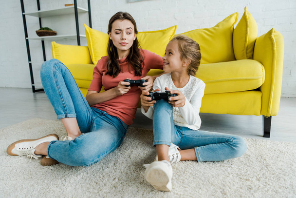 KYIV, UKRAINE - SEPTEMBER 4, 2019: babysitter and happy kid sitting on carpet and playing video game in living room  - Fotografie, Obrázek