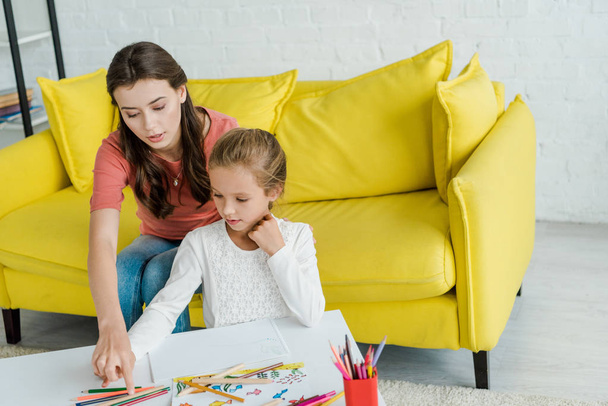 приваблива няня, що сидить на жовтому дивані біля дитини та кольорових олівцях у вітальні
  - Фото, зображення