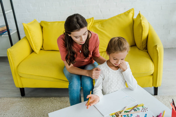 attractive babysitter sitting on yellow sofa near kid looking at drawn picture in living room  - Фото, изображение