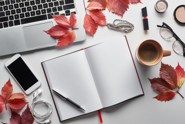 top view of laptop near smartphone, coffee cup, cosmetics, earphones, glasses, notebook and red leaves of wild grapes on white table - Photo, Image