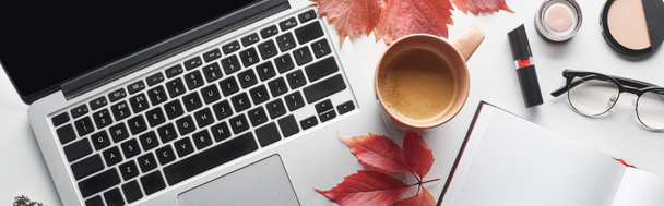 panoramische opname van laptop in de buurt van koffiebeker, notebook, glazen, cosmetica en rode bladeren van wilde druiven op witte tafel - Foto, afbeelding