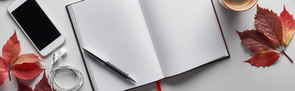 panoramiczne ujęcie pustego notebooka z długopisem, smartfonem, słuchawkami i czerwonymi liśćmi dzikich winogron na białym stole - Zdjęcie, obraz