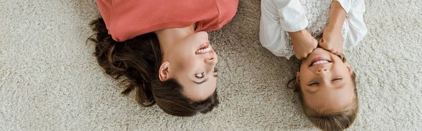 Panoramaaufnahme eines glücklichen Babysitters, der auf Teppich liegendes Kind betrachtet  - Foto, Bild