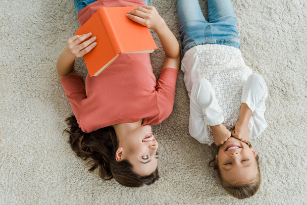 top view of happy babysitter holding book while looking at kid lying on carpet  - 写真・画像