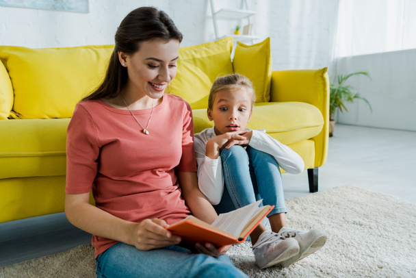 surprised kid sitting on carpet near happy babysitter with book  - Photo, Image