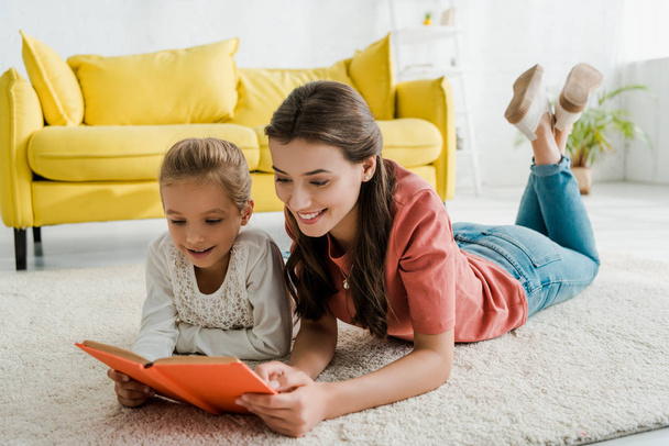 happy babysitter lying on carpet with cute kid while reading book  - Photo, Image