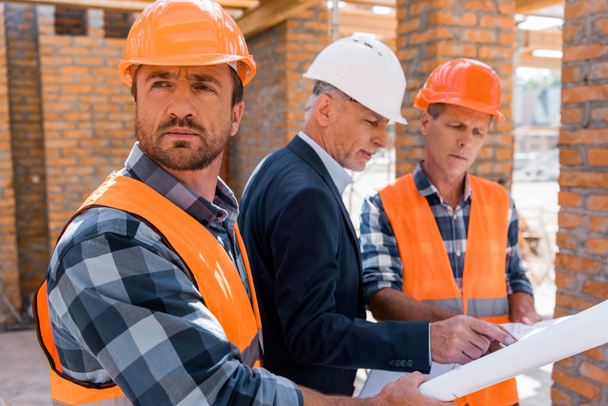 rakentajan valikoiva keskittyminen kypärään lähellä työtoveria ja liikemiestä
  - Valokuva, kuva