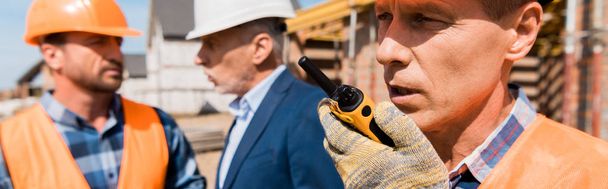 panoramic shot of builder holding walkie talkie while talking near coworker and businessman  - Photo, Image
