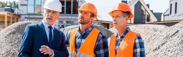 Bauarbeiter mit Helmen stehen neben Geschäftsleuten in Anzügen und Häusern  - Foto, Bild