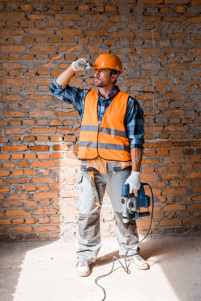 handsome bearded man holding hammer drill while touching helmet  - Zdjęcie, obraz