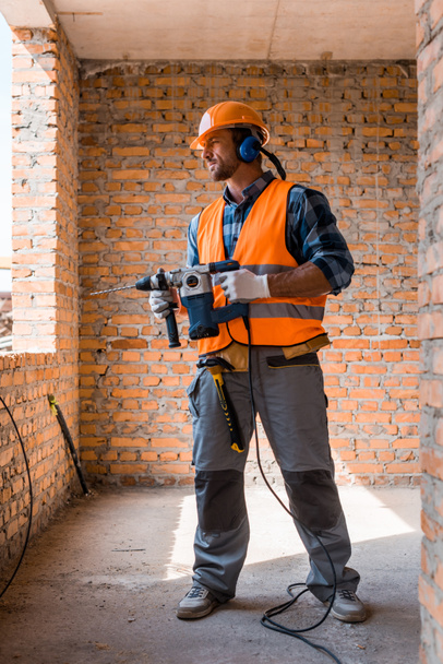 handsome bearded man holding hammer drill near brick walls  - Photo, Image
