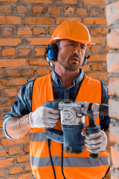 selective focus of handsome and bearded constructor holding hammer drill near brick wall  - Fotó, kép