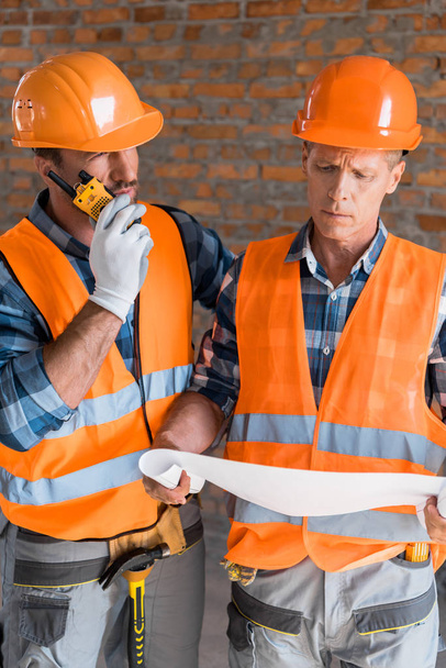 mature constructor holding blueprint near coworker with walkie talkie  - Foto, afbeelding