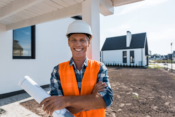 cheerful mature man holding blueprint and smiling near houses  - Fotografie, Obrázek