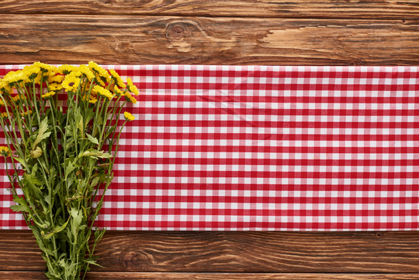 pohled shora na žluté květy na červeném kostkovaném ubrousku u dřevěného stolu - Fotografie, Obrázek