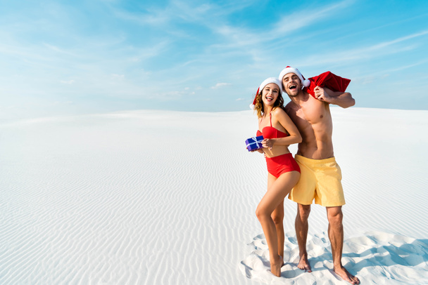 sexy and smiling girlfriend holding gift and boyfriend with santa sack on beach in Maldives  - Foto, afbeelding