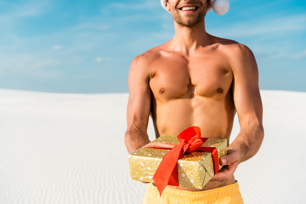 bijgesneden uitzicht van sexy man in santa hoed holding cadeau op het strand in Malediven  - Foto, afbeelding