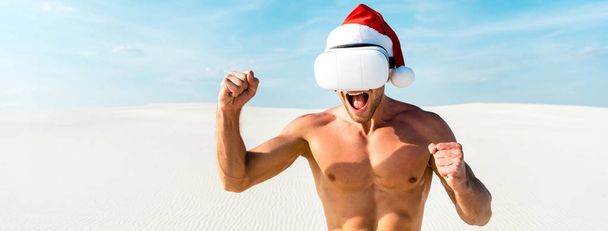 Sanal gerçeklik kulaklığıyla Noel Baba şapkalı seksi adamın Maldivler 'deki kumsalda evet jestini gösteren panoramik bir fotoğrafı.  - Fotoğraf, Görsel