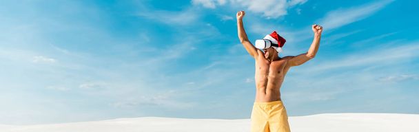 panoramic shot of sexy man in santa hat with virtual reality headset showing yes gesture on beach in Maldives  - Foto, afbeelding