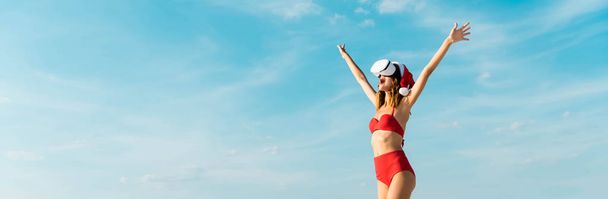 panoramic shot of sexy woman in santa hat and swimsuit with virtual reality headset on beach in Maldives  - 写真・画像