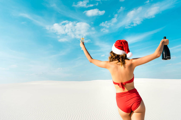 back view of sexy woman in santa hat and swimsuit holding champagne glass and bottle on beach in Maldives  - Foto, afbeelding